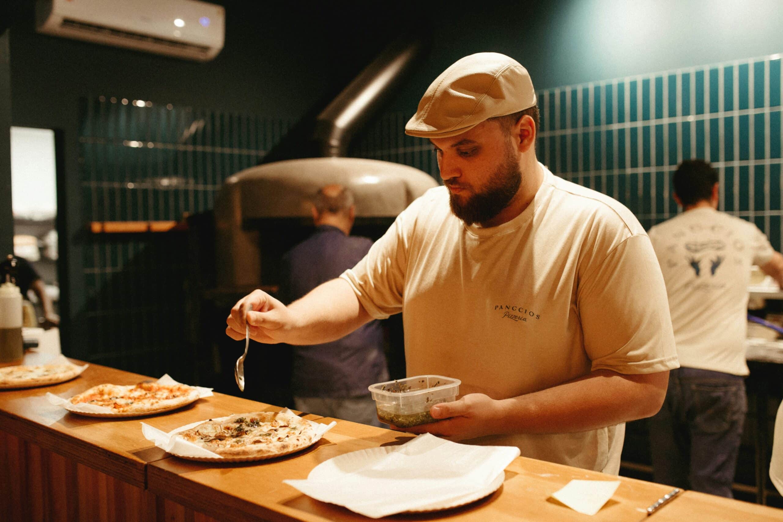 Chef Preparing Artisan Pizzas in a Rustic Kitchen Bearded chef adding toppings to pizza in a traditional pizzeria kitchen