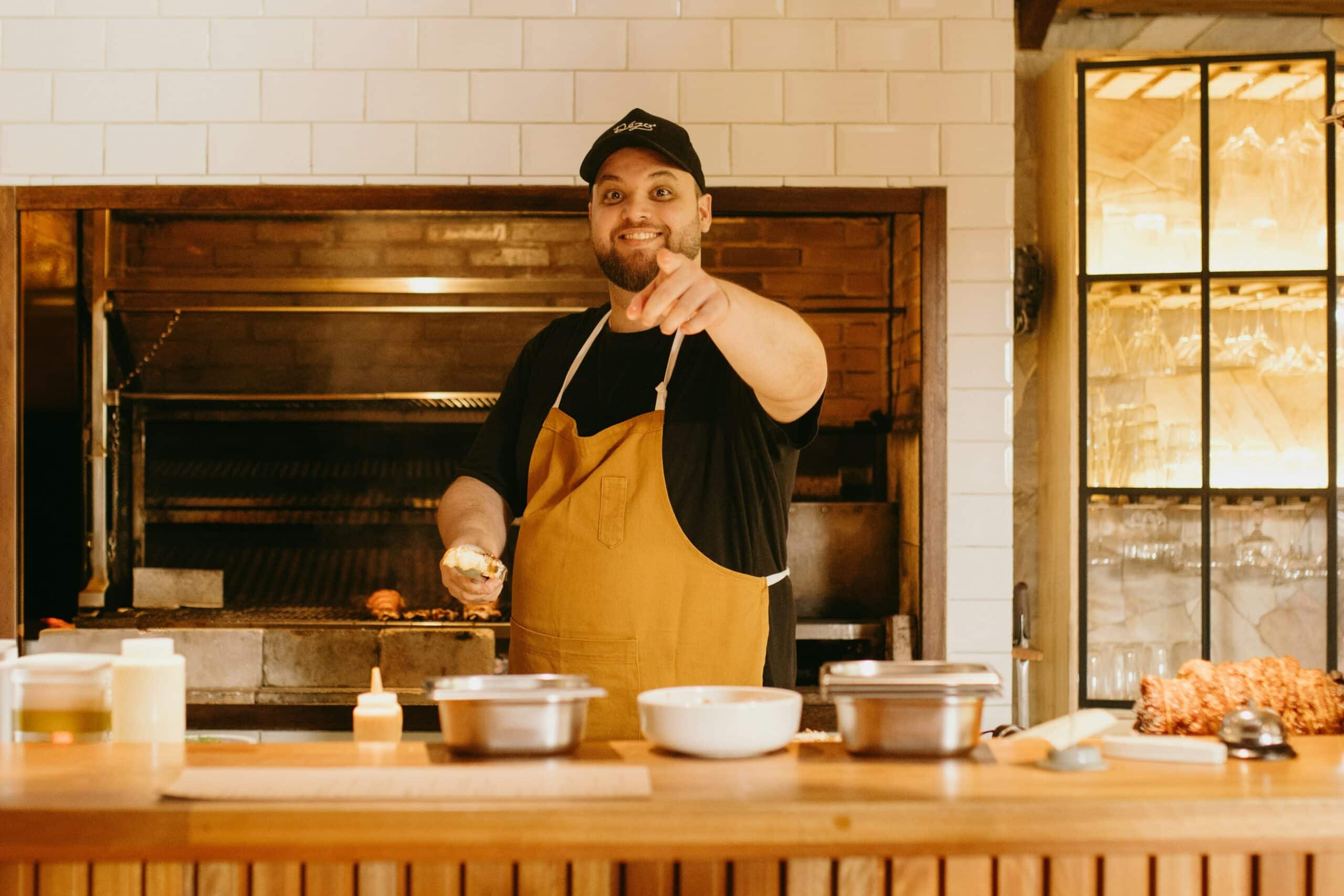 Smiling Chef in Apron Pointing from a Cozy Kitchen Happy chef wearing a yellow apron, pointing and smiling in a warm kitchen