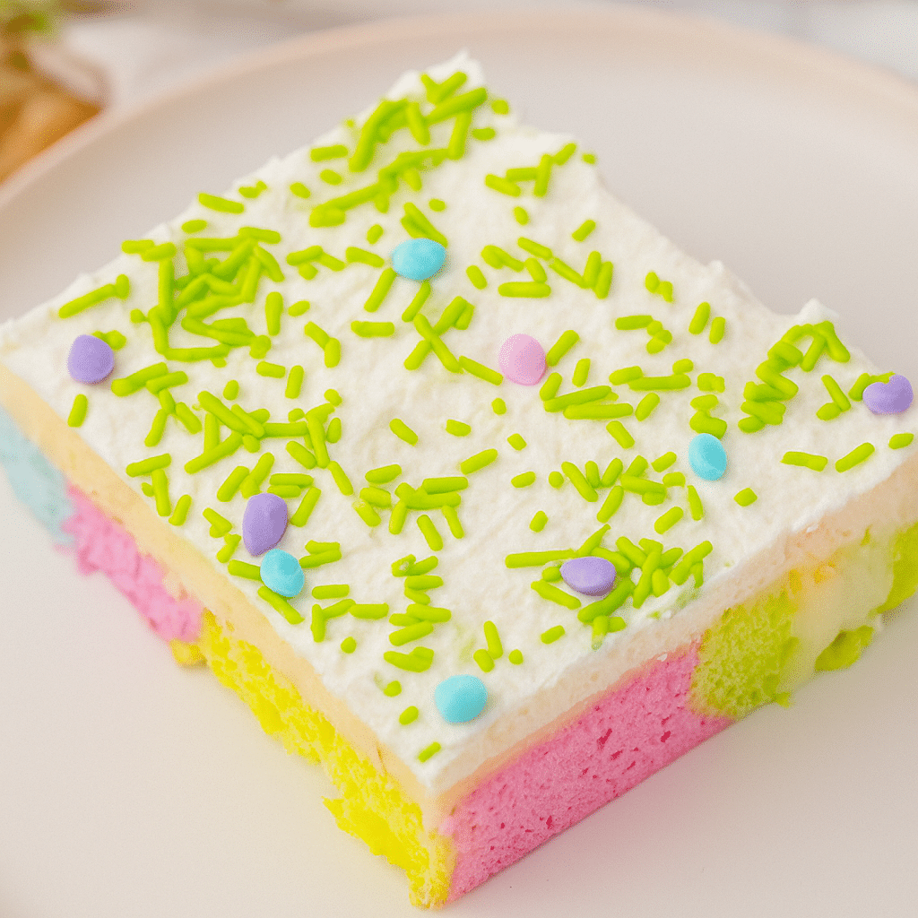 Easter Poke Cake Slice | Bright, Moist & Colorful Dessert A colorful slice of Easter poke cake with green sprinkles and pastel layers of pink, yellow, and blue, topped with whipped cream.