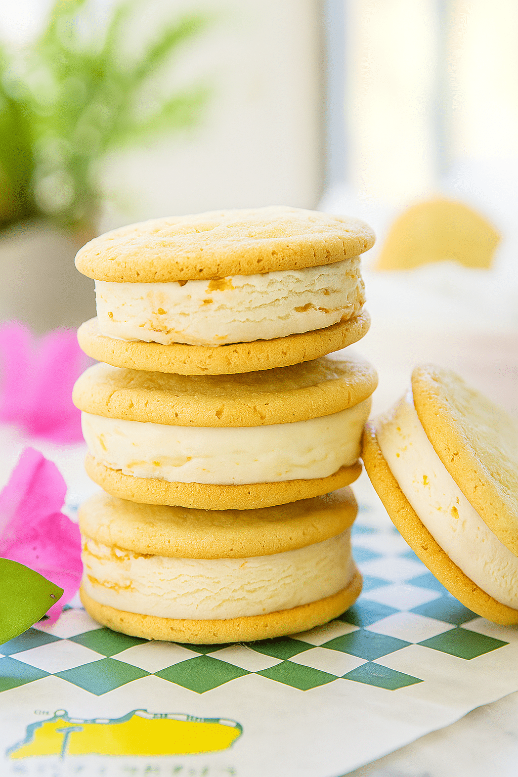 Homemade Ice Cream Sandwich Cookies | Easy, Creamy, and Delicious A close-up image of three stacked vanilla Taste Of The Masters: Make Peach Ice Cream Sandwiches cookies with creamy filling, placed on a checkered napkin, surrounded by flowers in soft light.