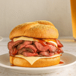 Homemade Roast Beef Sandwich Fats Lat's Make It Now 3 Close-up of a roast beef sandwich stacked with tender beef slices, melted cheese, and barbecue sauce on a toasted cornmeal bun, served on a white plate.
