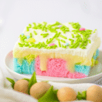 Easter Poke Cake Recipe: The Most Colorful & Easy Dessert 3 A square slice of pastel-colored Easter cake topped with creamy frosting and green sprinkles, sitting on a white plate.