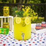 Pickled Lemonade: A Bold, Crisp Flavor Surprise, Try now 3 A large glass jar filled with pickle lemonade garnished with dill and lemon slices, displayed on a star-patterned table outdoors with a blurred background.